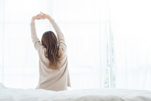 Opinión trasera la mujer asiática joven hermosa feliz que despierta por la mañana, sentándose en cama