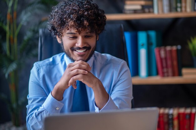 Optimismo. Joven barbudo con cabello rizado oscuro sosteniendo sus manos cerca de la barba sentado en la oficina en la mesa sonriendo mirando la pantalla del portátil