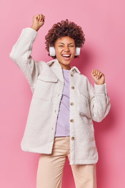 Optimista, alegre, despreocupada, joven baila con triunfo tiene un estado de ánimo optimista, usa una chaqueta elegante y pantalones, escucha música favorita en auriculares, se mueve contra la pared rosa