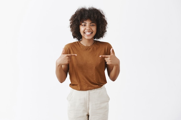 optimista amigable chica afroamericana con cabello rizado que quiere ser elegida sugiriéndose como candidata apuntando al pecho y sonriendo alegremente con expresión feliz