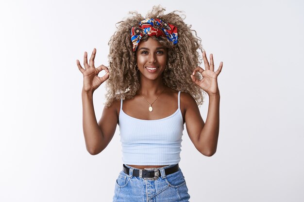 Optimista atractiva chica rubia de pelo rizado con peinado afro, diadema, muestra ok, ok signo de confirmación y sonriendo, asentir con aceptación, como una idea increíble, dar aprobación