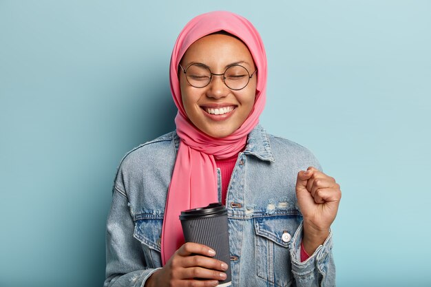 Optimista encantadora mujer musulmana aprieta el puño, cierra los ojos de placer