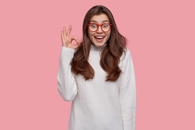 Optimista mujer joven de pelo oscuro muestra cero signos o gesto bien, sonríe ampliamente, viste un suéter blanco, aprueba todo lo bueno