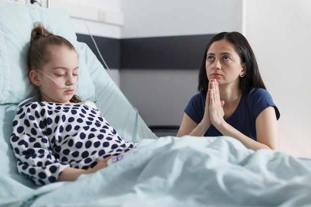 Orando madre preocupada sentada en la sala de la clínica pediátrica mientras una pequeña hija enferma duerme en la cama del hospital. Niña malsana descansando mientras una mujer triste y pensativa reza por su salud.