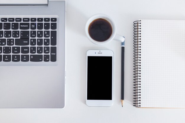 Ordenador portátil, teléfono móvil, café, lápiz y cuaderno espiral sobre el escritorio blanco