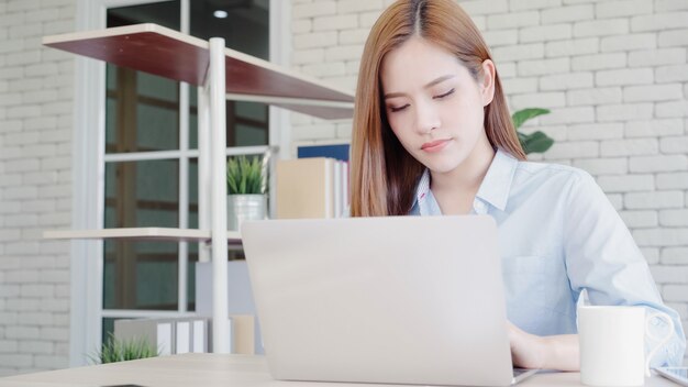 Ordenador portátil de trabajo sonriente de la mujer asiática joven hermosa en el escritorio en sala de estar en casa.