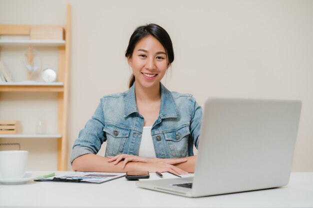 Ordenador portátil de trabajo sonriente de la mujer asiática joven hermosa en el escritorio en sala de estar en casa.