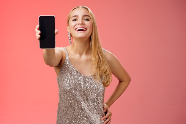 Orgullosa alegre encantadora alegre rubia europea con elegante vestido plateado brillante mantenga la cintura de la mano confiada extender el brazo mostrando la pantalla del teléfono inteligente presente nuevo dispositivo de aplicación impresionante, fondo rojo.