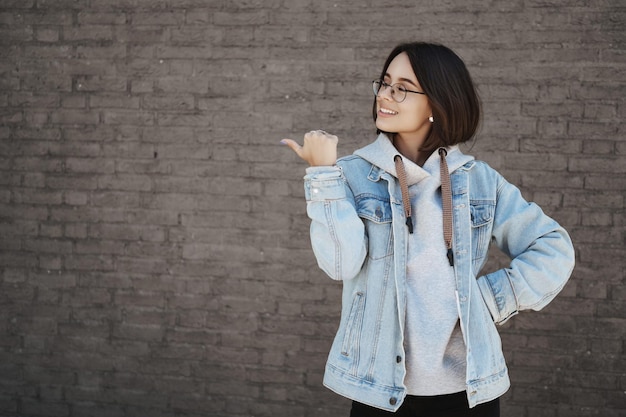 Orgullosa y feliz chica caucásica sonriente con el pelo corto y gafas que le muestra la pancarta de la empresa promocional o un anuncio que apunta al espacio libre de la pared de ladrillos para su logotipo recomendando