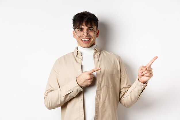 Orgulloso y feliz joven con gafas mostrando el logo, señalando con el dedo en la esquina superior derecha y sonriendo, de pie sobre fondo blanco.