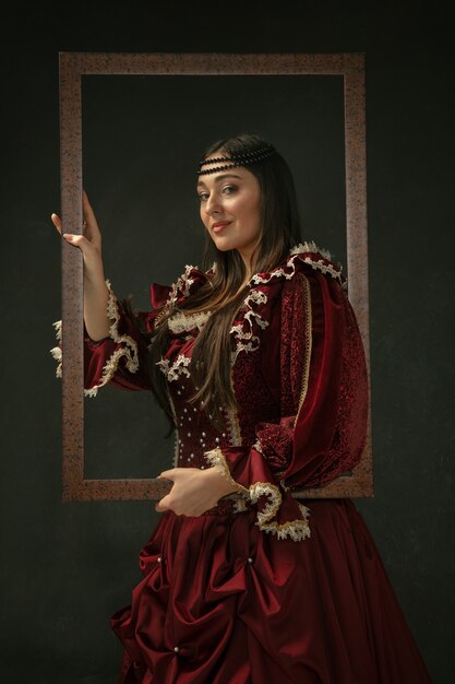 Orgulloso. Retrato de mujer joven medieval en ropa vintage roja de pie sobre fondo oscuro. Modelo femenino como duquesa, persona real. Concepto de comparación de épocas, moderno, moda, belleza.