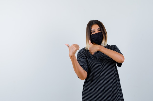 Foto gratuita ortrait de dama apuntando hacia la izquierda con los pulgares en vestido negro, máscara médica y mirando confiada vista frontal