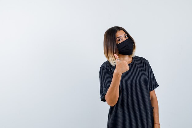 Ortrait de dama mostrando el pulgar hacia arriba en vestido negro, máscara médica y mirando confiada vista frontal