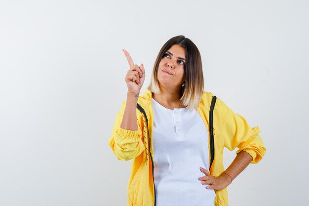 Ortrait de mujer apuntando hacia arriba en camiseta, chaqueta y mirando esperanzada vista frontal