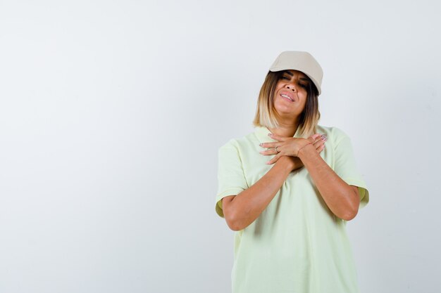 Ortrait de señorita cogidos de la mano en el pecho en camiseta, gorra y mirando alegre vista frontal