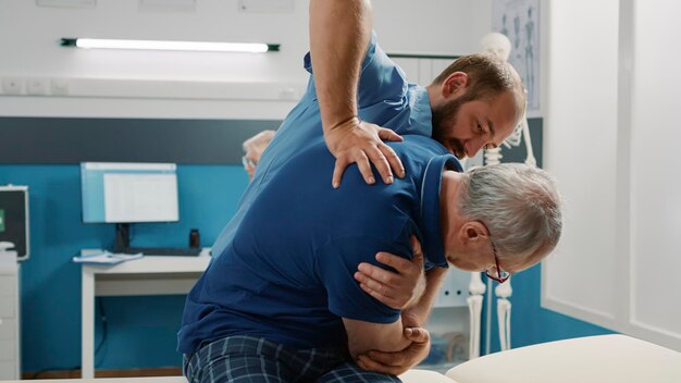 Osteópata ayudando a un anciano a romper huesos en el gabinete, haciendo ejercicio de fisioterapia. Asistente masculino que utiliza un procedimiento de fisioterapia para aumentar la movilidad y tratar al paciente.