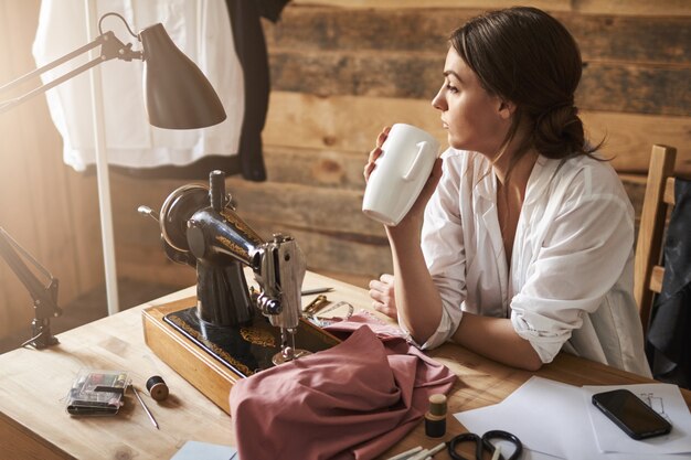 Otro día en el taller llegó a su fin. Soñadora alcantarilla femenina pensativa mirando a un lado mientras está sentado cerca de la máquina de coser, tomando té y tomando un descanso del trabajo. Diseñador recarga con café caliente