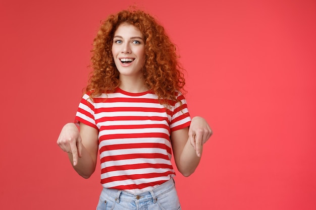 Oye, compruébalo amigo. Descarado atractivo pelirrojo descarado novia elegante peinado rizado usar camiseta de verano apuntando hacia abajo con el dedo índice sonriendo divertido encantado presente impresionante recomendación.