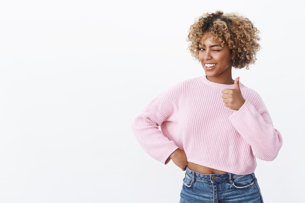 Oye, eres el mejor. Retrato de mujer afroamericana carismática coqueta y feliz con corte de pelo rubio guiñando un ojo con alegría y sonriendo mientras muestra el pulgar hacia arriba en gesto de aprobación y aprobación sobre la pared blanca