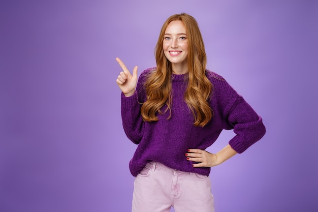 Oye, haz clic aquí. Retrato de mujer pelirroja encantadora y linda de aspecto amistoso en suéter púrpura apuntando a la esquina superior izquierda con el dedo índice y sonriendo, mostrando espacio de copia, recomendando, ofreciendo.