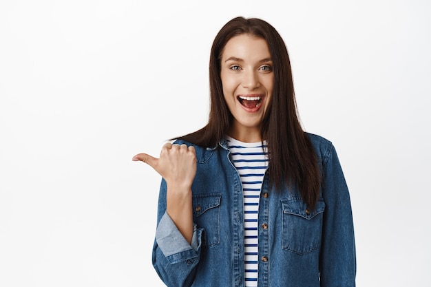 Oye, ve por ese camino. Mujer amigable sonriente sugiere seguir esta página, señalar con el dedo a la izquierda y lucir emocionada, invitar a la gente, recomendar artículo, fondo blanco