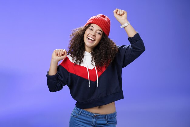 Oye, ven a bailar conmigo. Mujer alegre brillante y elegante de aspecto amistoso con peinado rizado con gorro cálido levantando las manos como disfrutando de un gran día, divirtiéndose al aire libre sobre una pared azul.