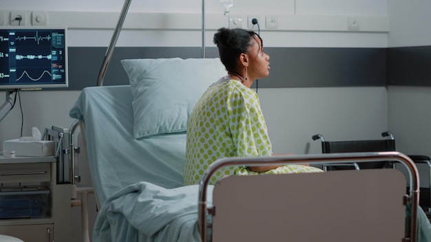 Paciente afroamericano con enfermedad sentado en la cama de la sala del hospital, esperando recibir tratamiento y asistencia médica del médico. Mujer joven con bolsa de goteo intravenoso y monitor de frecuencia cardíaca