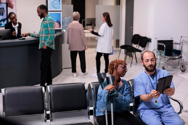 Paciente afroamericano y enfermera mirando una tableta para encontrar un diagnóstico de enfermedad en un dispositivo digital, que sufre de discapacidad física en el área de espera. Mujer y especialista en el vestíbulo del hospital.