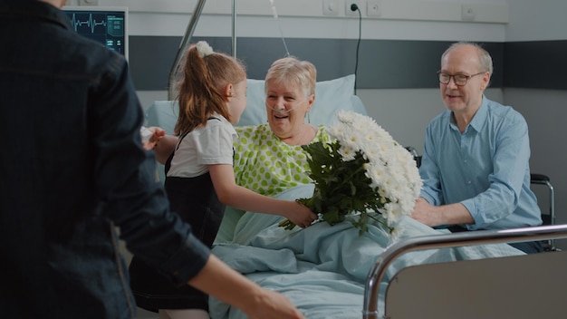 Paciente anciano que recibe la visita del niño y la madre en la sala del hospital. Niño con flores corriendo para abrazar a la mujer enferma en la cama, visitando a la abuela para consolarla y ayudarla a recuperarse. Familia en la clínica