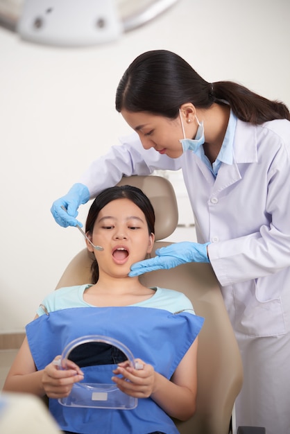 Paciente asiático sentado con la boca abierta y sosteniendo el espejo, y el dentista revisando sus dientes