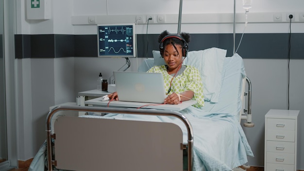 Paciente con enfermedad que usa auriculares para escuchar música mientras está sentado en la cama de la sala de hospital. Mujer joven mirando portátil y escuchando sonidos de audio en auriculares. Adulto con enfermedad