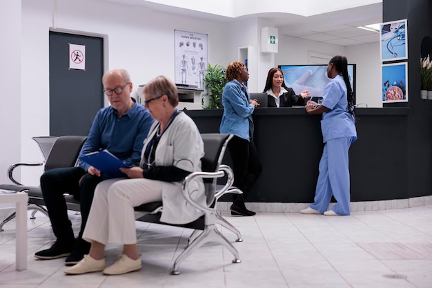 Paciente y enfermera sentados en el mostrador de recepción, hablando con una recepcionista sobre el diagnóstico de enfermedades y el apoyo sanitario. Diversas personas que trabajan en el centro de salud en el mostrador de registro.
