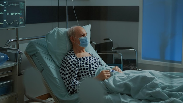 Paciente enfermo sentado en la cama de la sala de hospital con mascarilla y oxímetro en la clínica. Anciano con enfermedad esperando tratamiento médico en bolsa de goteo intravenoso para curar enfermedades y problemas de salud