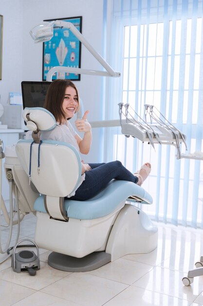 Paciente femenino sonriente que se sienta en la silla que muestra el pulgar arriba en clínica dental