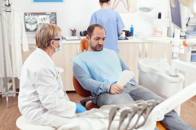 Paciente hombre mirando en el espejo después del equipo médico estomatológico finalizando la cirugía dental durante el procedimiento de estomatología