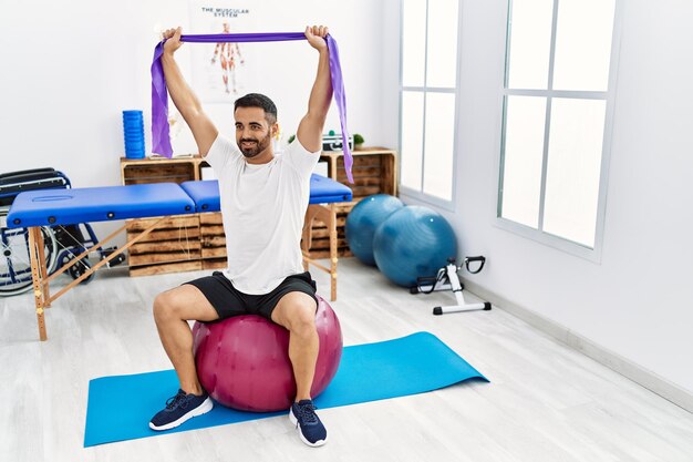 Paciente joven hispano sonriendo confiado teniendo sesión de rehabilitación usando fit ball y banda elástica en la clínica