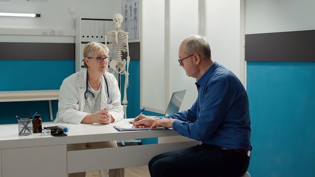 Paciente jubilado que firma el documento de cita para recibir tratamiento con receta y apoyo del sistema de atención médica. Doctor consultando a un enfermo en un gabinete médico, firma en documentos.