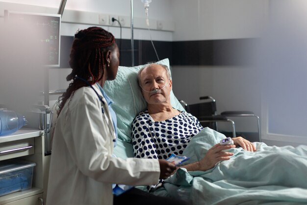 Paciente mayor enfermo acostado en la cama respirando a través de un tubo de ensayo de oxígeno, escuchando a su médico