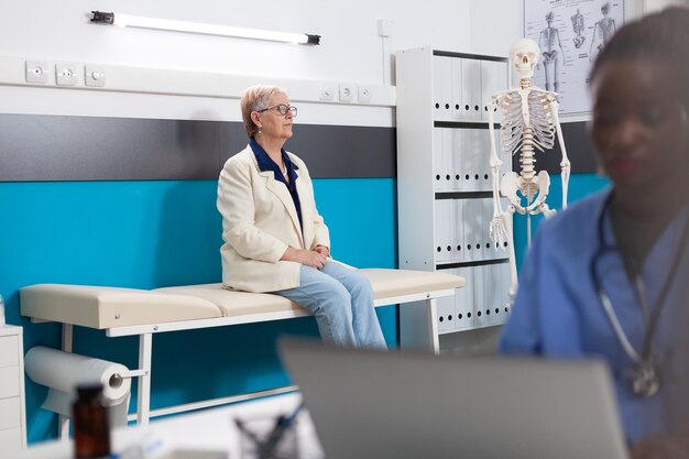 Paciente mujer senior pensionista de pie en la cama esperando la experiencia de la enfermedad durante la cita médica en la oficina del hospital. Enfermera afroamericana escribiendo tratamiento con medicamentos. Asistencia de soporte