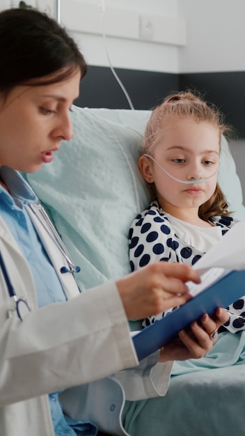 Paciente niño enfermo con tubo nasal de oxígeno descansando en la cama recuperándose después de la cirugía de enfermedad respiratoria durante la consulta médica. Médico mujer médico analizando la experiencia de la enfermedad en la sala del hospital