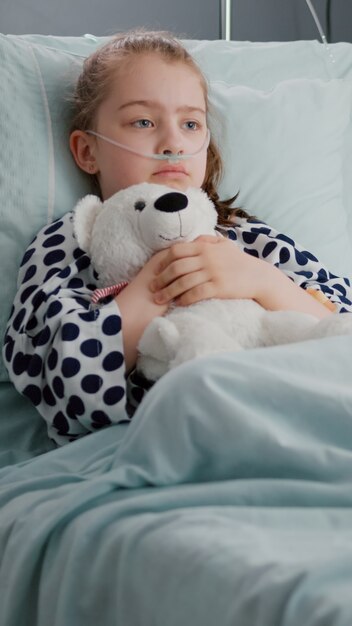 Paciente niño preocupado solo con tubo nasal de oxígeno descansando en la cama sosteniendo oso teedy