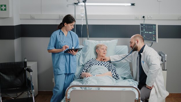 Paciente con tubo de oxígeno nasal que recibe consulta del médico con estetoscopio para examen de los latidos del corazón. Médico que consulta a una mujer jubilada con enfermedad mientras la enfermera ayuda con la tableta