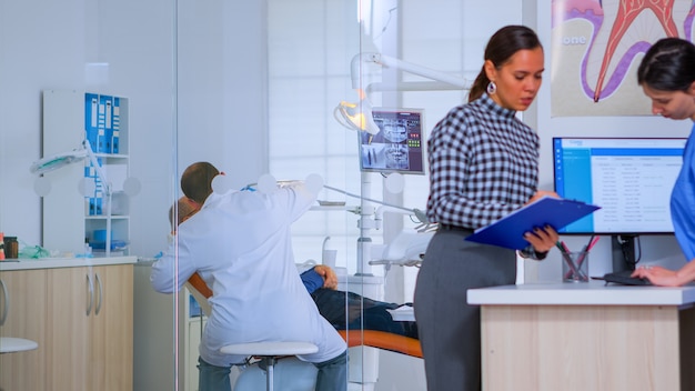 Pacientes que solicitan ayuda para completar el formulario de registro dental preparándose para la ejecución. Mujer mayor sentada en una silla en la sala de espera de la concurrida oficina del ortodoncista mientras el médico trabaja en segundo plano.