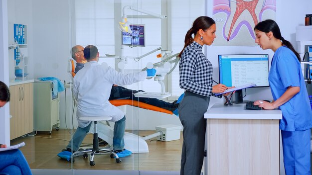 Pacientes que solicitan información cumplimentando el documento dental preparándose para la ejecución de los dientes. Mujer mayor sentada en una silla en la sala de espera de la concurrida oficina del ortodoncista mientras el médico trabaja en segundo plano.