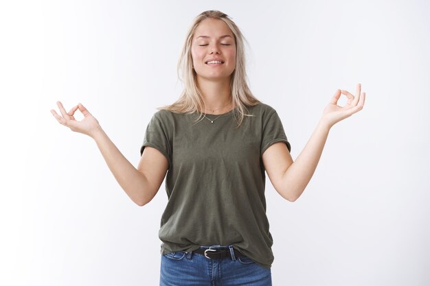 Pacífica joven atractiva mujer de 25 años con cabello rubio inhalando aire fresco con los ojos cerrados y sonriendo aliviado y feliz meditando en posición de loto con signos de mudra hacia los lados contra el fondo blanco.