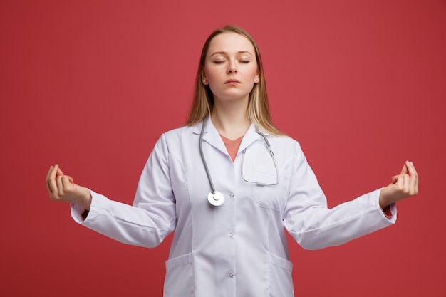 Pacífica joven doctora rubia vistiendo bata médica y un estetoscopio alrededor del cuello meditando con los ojos cerrados