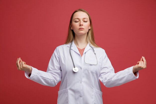 Pacífica joven doctora rubia vistiendo bata médica y un estetoscopio alrededor del cuello meditando con los ojos cerrados