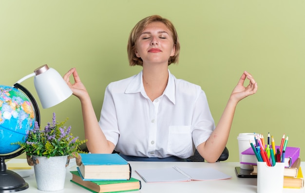 Pacífica joven estudiante rubia sentada en un escritorio con herramientas escolares meditando con los ojos cerrados aislado en la pared verde oliva