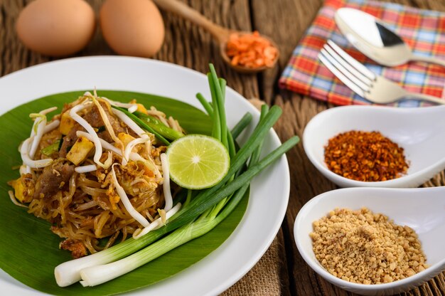 Pad Thai en un plato blanco con limón, huevos y condimentos en una mesa de madera.