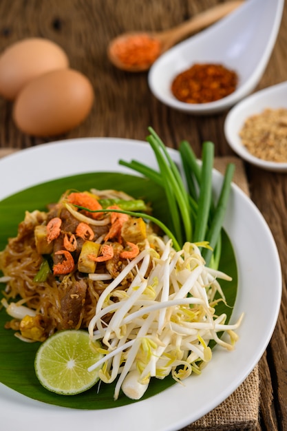 Pad Thai en un plato blanco con limón, huevos y condimentos en una mesa de madera.